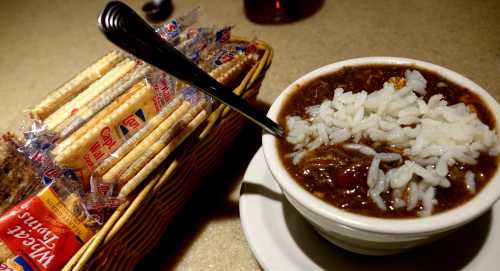 A bowl of chili topped with rice, accompanied by a basket of crackers on a table.