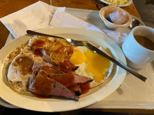 A plate of breakfast featuring eggs, hash browns, sliced meat, a biscuit, and a cup of coffee on the side.