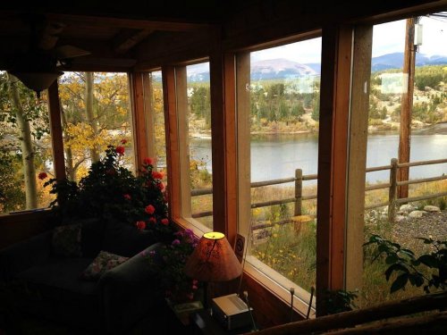 Cozy interior with a sofa and lamp, framed by large windows overlooking a serene lake and autumn foliage.
