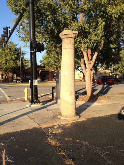 A weathered stone column stands at a street corner, surrounded by trees and a traffic signal.