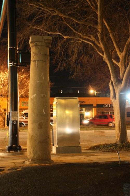 A weathered stone column stands beside a metallic utility box at night, with trees and storefronts in the background.