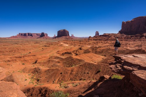 A person stands on a rocky ledge overlooking a vast desert landscape with red rock formations under a clear blue sky.