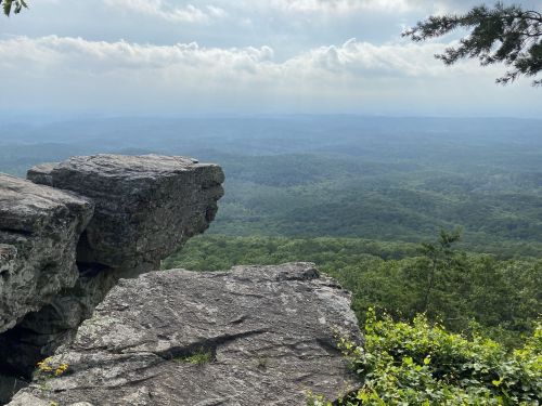 A rocky outcrop overlooks a vast green valley under a cloudy sky, with distant mountains in the background.