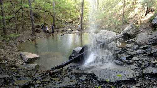 A serene waterfall cascades into a pond surrounded by rocks and trees, with people enjoying the natural setting nearby.