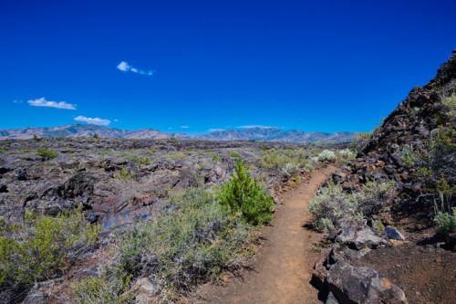 A winding dirt path through rocky terrain under a clear blue sky, with distant mountains in the background.