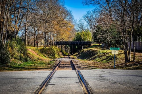 A quiet scene of a railway crossing with a bridge overhead, surrounded by trees and a clear blue sky.
