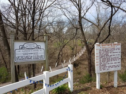 A sign for the Swinging Bridge in Louisa County, with a bridge visible in the background and a historical information board.