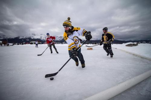 Three players skate on an outdoor ice rink, playing hockey against a backdrop of cloudy mountains.