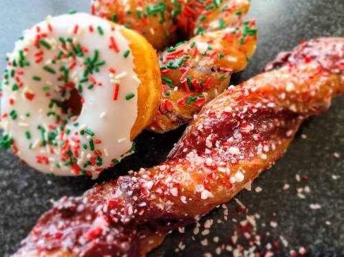 A festive assortment of donuts topped with icing and sprinkles, alongside a twisted pastry with red filling and sugar.