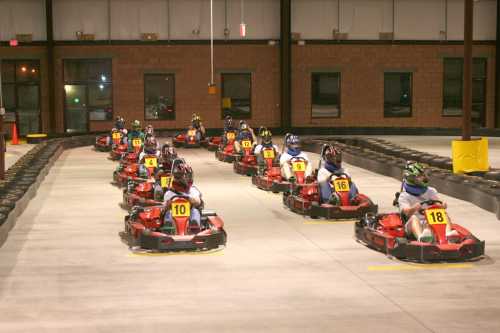 A group of go-karts racing on an indoor track, with drivers wearing helmets and numbered vests.