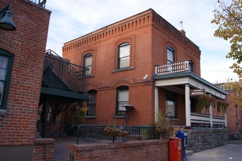 Historic brick hotel building with a porch, stairs, and a sign, surrounded by trees and a blue sky.