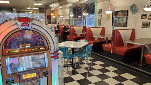 A retro diner interior featuring a jukebox, red booths, and a checkered floor, with a customer seated in the background.