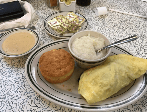 A plate with a wrapped omelet, grits, a biscuit, and a bowl of gravy on a patterned tablecloth.
