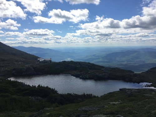 A scenic view of a mountain landscape with a lake, rolling hills, and a cloudy blue sky in the background.