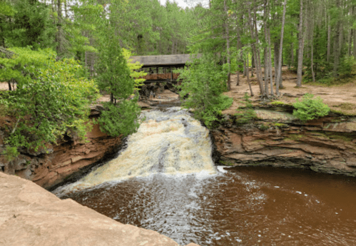 A serene waterfall flows into a brown river, surrounded by lush green trees and a wooden bridge in the background.