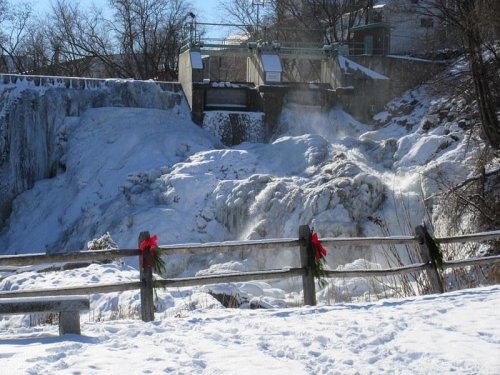 Frozen waterfall with snow-covered rocks, a wooden fence adorned with red bows, and trees in the background.