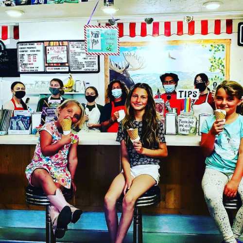 A group of children and staff at an ice cream shop, smiling and holding ice cream cones, with a colorful backdrop.