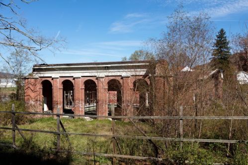 Abandoned brick building with arched windows, surrounded by overgrown vegetation and a wooden fence under a clear blue sky.