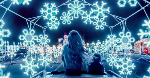 A child and an adult sit in a vehicle, surrounded by glowing snowflake lights in a festive display at night.