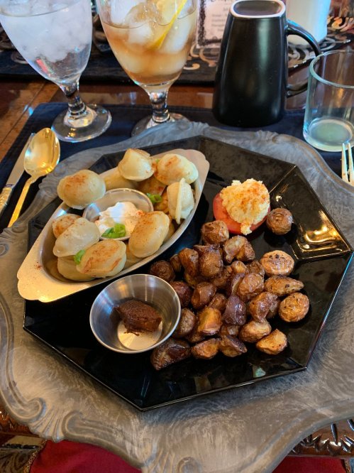 A black plate with assorted appetizers: stuffed dumplings, roasted potatoes, and a small bowl of sauce. Drinks in the background.