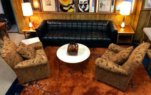 A cozy living room setup featuring a black leather sofa, two patterned armchairs, a round coffee table, and warm lighting.