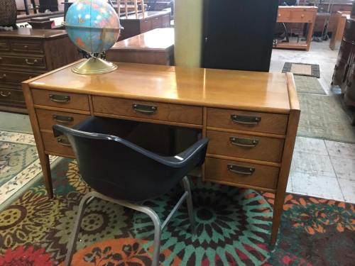 A wooden desk with multiple drawers and a globe on top, accompanied by a blue chair, set in a furniture store.