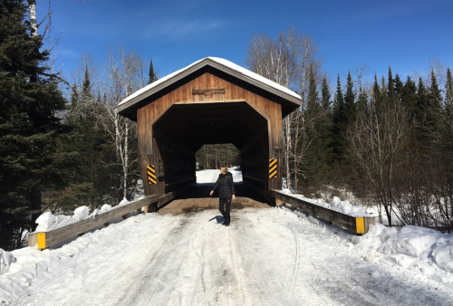 A person walks towards a wooden covered bridge surrounded by snow and trees under a clear blue sky.