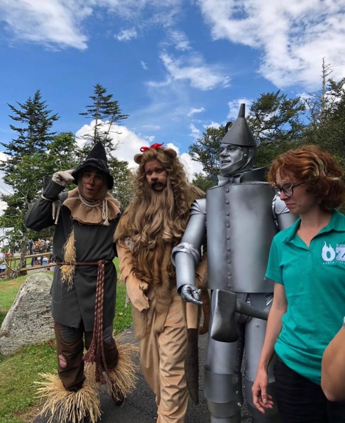Three people dressed as characters from "The Wizard of Oz" walk together outdoors, with trees and a blue sky in the background.