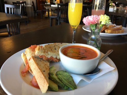 A plate with a grilled cheese sandwich, tomato soup, avocado slices, and a glass of mimosa on a restaurant table.