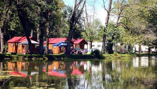 A serene lakeside scene with wooden cabins and trees reflecting in the water under a clear blue sky.