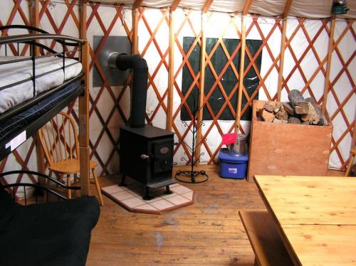 Interior of a yurt featuring a wood stove, bunk bed, wooden table, and stacked firewood against a wall.