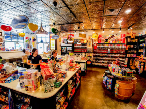 A colorful retro store interior with two cashiers, shelves filled with snacks, drinks, and vintage decor.