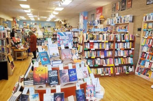 A cozy bookstore interior with shelves of books, a display table, and a customer browsing in the background.