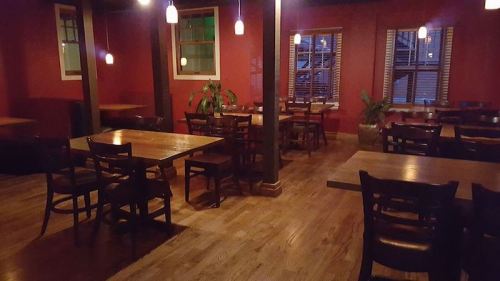 A dimly lit restaurant interior with wooden tables, chairs, and red walls, featuring plants and pendant lighting.