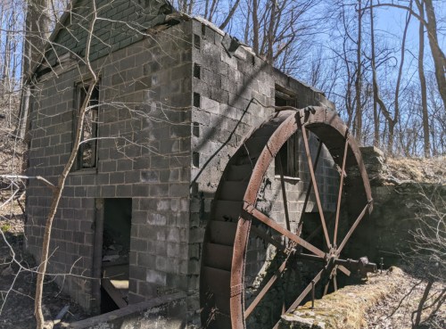 Abandoned building with a rusted water wheel, surrounded by trees and underbrush on a sunny day.