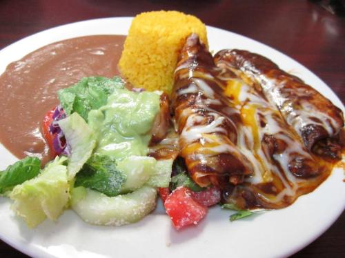 A plate of enchiladas topped with cheese, served with rice, refried beans, and a fresh salad.