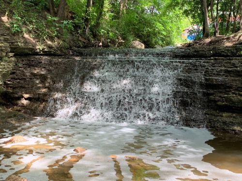 A serene waterfall cascading over rocky ledges, surrounded by lush greenery and a calm pool below.