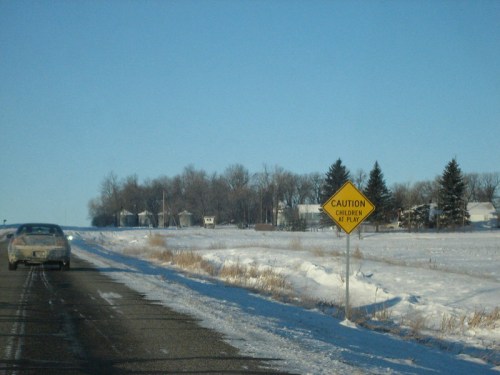 A snowy road with a "Caution: Children at Play" sign and a car driving past, surrounded by trees and houses.