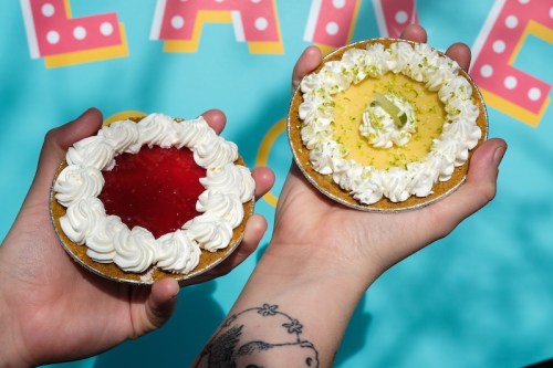 Two hands hold colorful desserts: one with red jelly and whipped cream, the other with lime filling and decorative cream.
