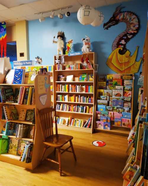 A cozy children's bookstore with colorful shelves, a wooden chair, and whimsical decorations on the walls.