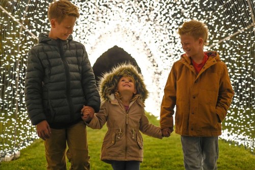 Two boys and a girl walk hand in hand through a tunnel of sparkling lights, smiling and enjoying the moment.