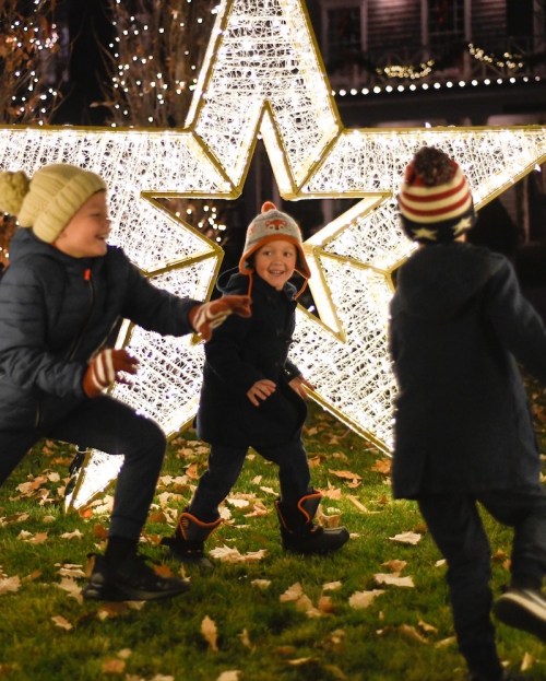Three children play joyfully in front of a large illuminated star decoration, surrounded by fallen leaves.