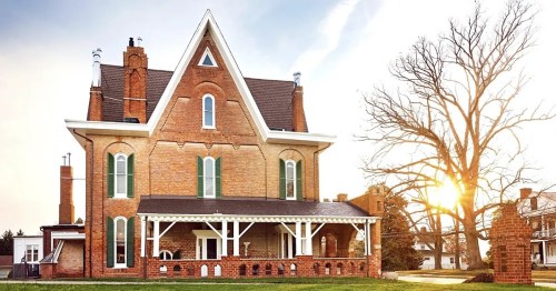 A large, historic brick house with a gabled roof, surrounded by a grassy area and a tree, illuminated by sunset.