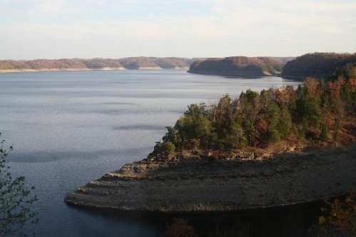 A serene lake surrounded by wooded hills, with calm waters reflecting the sky and autumn foliage.