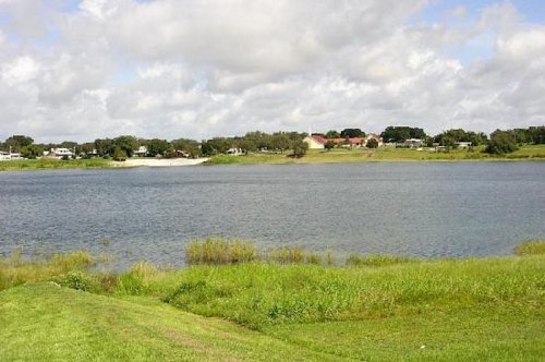 A serene lake surrounded by green grass and trees, with a cloudy sky and houses in the background.