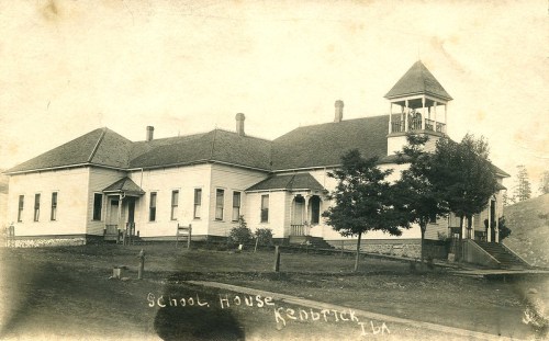 Historic schoolhouse with a bell tower, surrounded by trees, in a rural setting. Black and white photo.