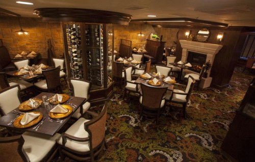 Elegant restaurant interior with wooden furniture, neatly set tables, a wine display, and a cozy fireplace.
