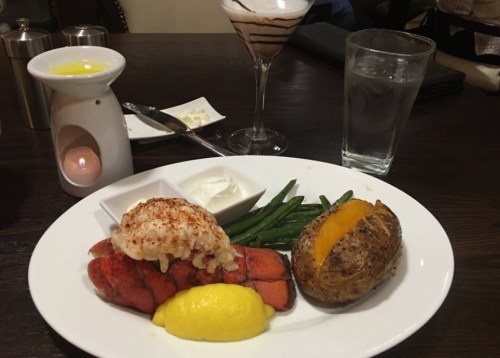 A plate with lobster, lemon, green beans, a baked potato, and a dessert drink on a wooden table.