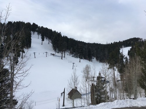Snow-covered ski slope with chairlifts and pine trees under a cloudy sky.