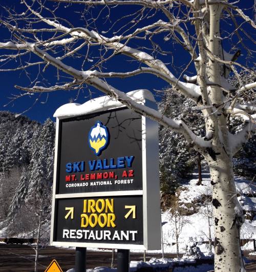Sign for Ski Valley and Iron Door Restaurant, surrounded by snow-covered trees and mountains under a clear blue sky.